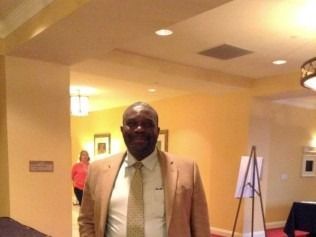 Man in a blazer and tie in a hallway with a woman in the background. Beige walls, warm lighting.
