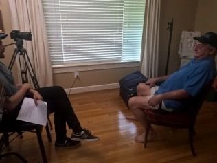 Two men in a room; one interviews the other. A camera records the interview. 