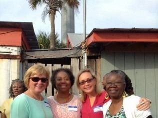 Four women smiling, posing outdoors by buildings, a palm tree and a tall structure in the background.