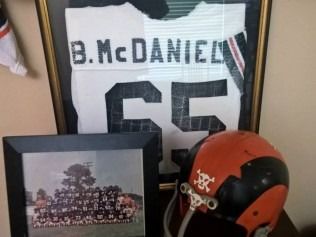 Framed football jersey, team photo, and orange helmet with skull, on a shelf.