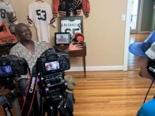 Man being interviewed with cameras; sports memorabilia background.