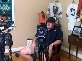 Man seated, speaking into a camera on a tripod. 