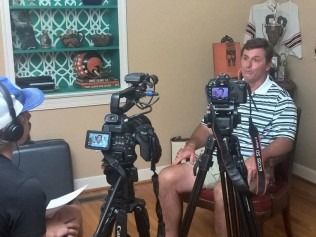 Man in striped shirt being interviewed, two cameras on tripods.