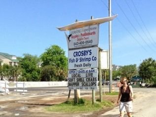 Sign for Crosby's Fish & Shrimp Co. with phone number, woman stands nearby, clear sky in background.