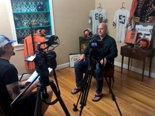 Man being interviewed in room with football memorabilia; cameras, jerseys, and a helmet are visible.