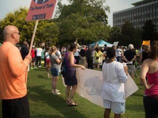 Group of people protesting in a park, holding signs. One sign reads