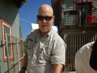 Bald man wearing sunglasses and a tan shirt, standing outside near buildings.