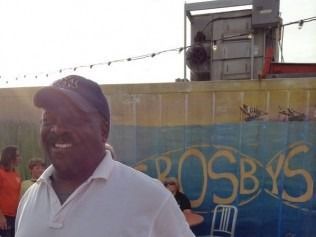 Man in a white shirt and baseball cap smiles outdoors; backdrop has Crosby's sign and machinery.