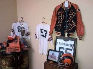 Sports memorabilia display: football jerseys, jacket, helmet, trophy, framed items, and a drum-like table.