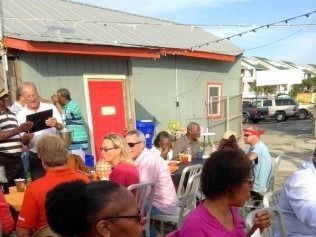 People seated at outdoor tables outside a building, socializing and eating, with cars parked nearby.