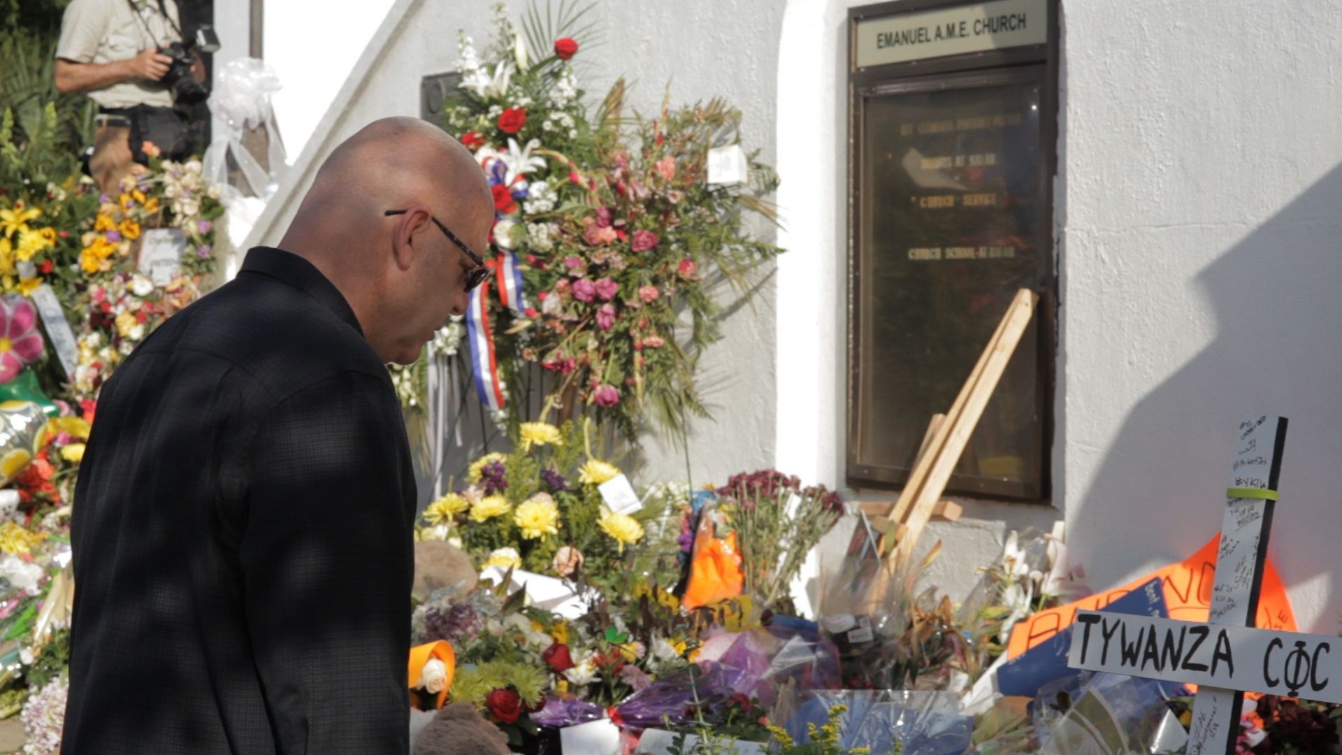 Man in black suit looking at a memorial with flowers and a plaque.