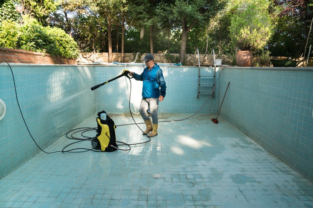 Man pressure washes an empty, light blue tiled swimming pool outdoors.