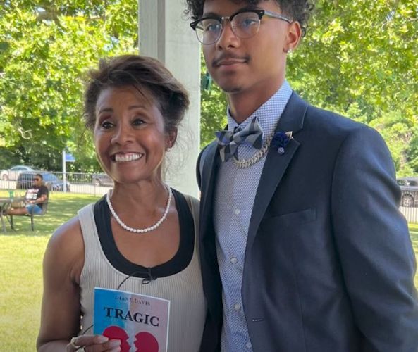 Diane standing next her grandson posing for a picture while the woman is holding a book titled Tragic Victory.