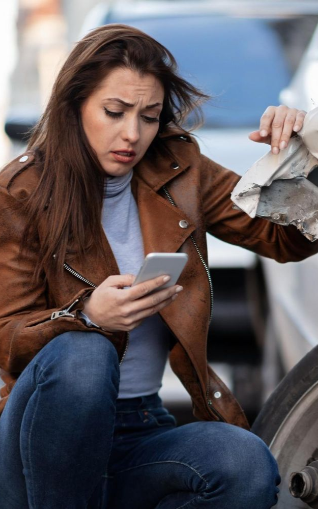 Woman in brown jacket, distressed, looking at phone near damaged car tire.