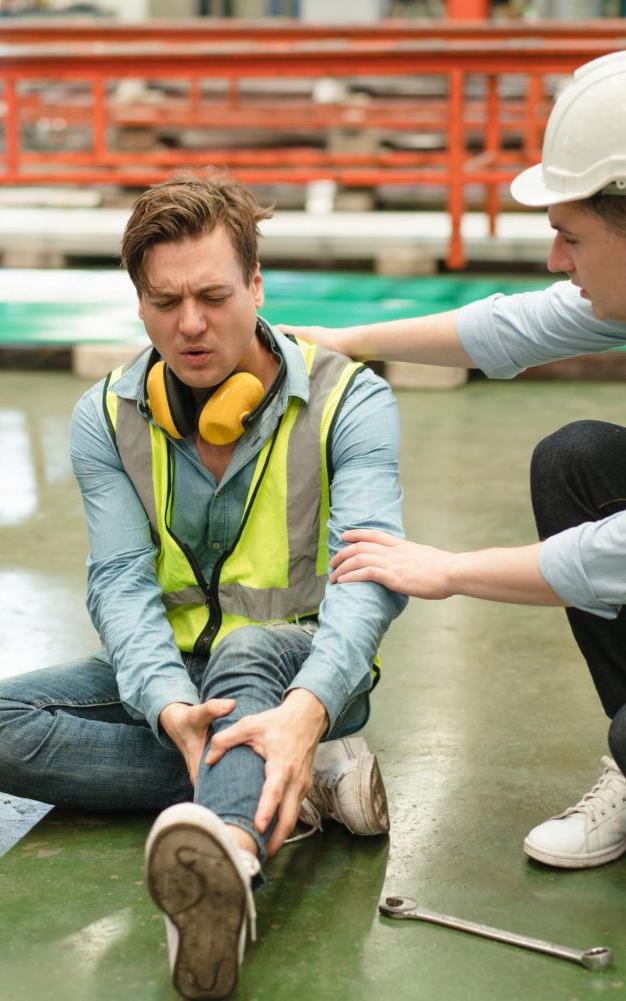 Man injured on factory floor, clutching leg, being helped by a woman.