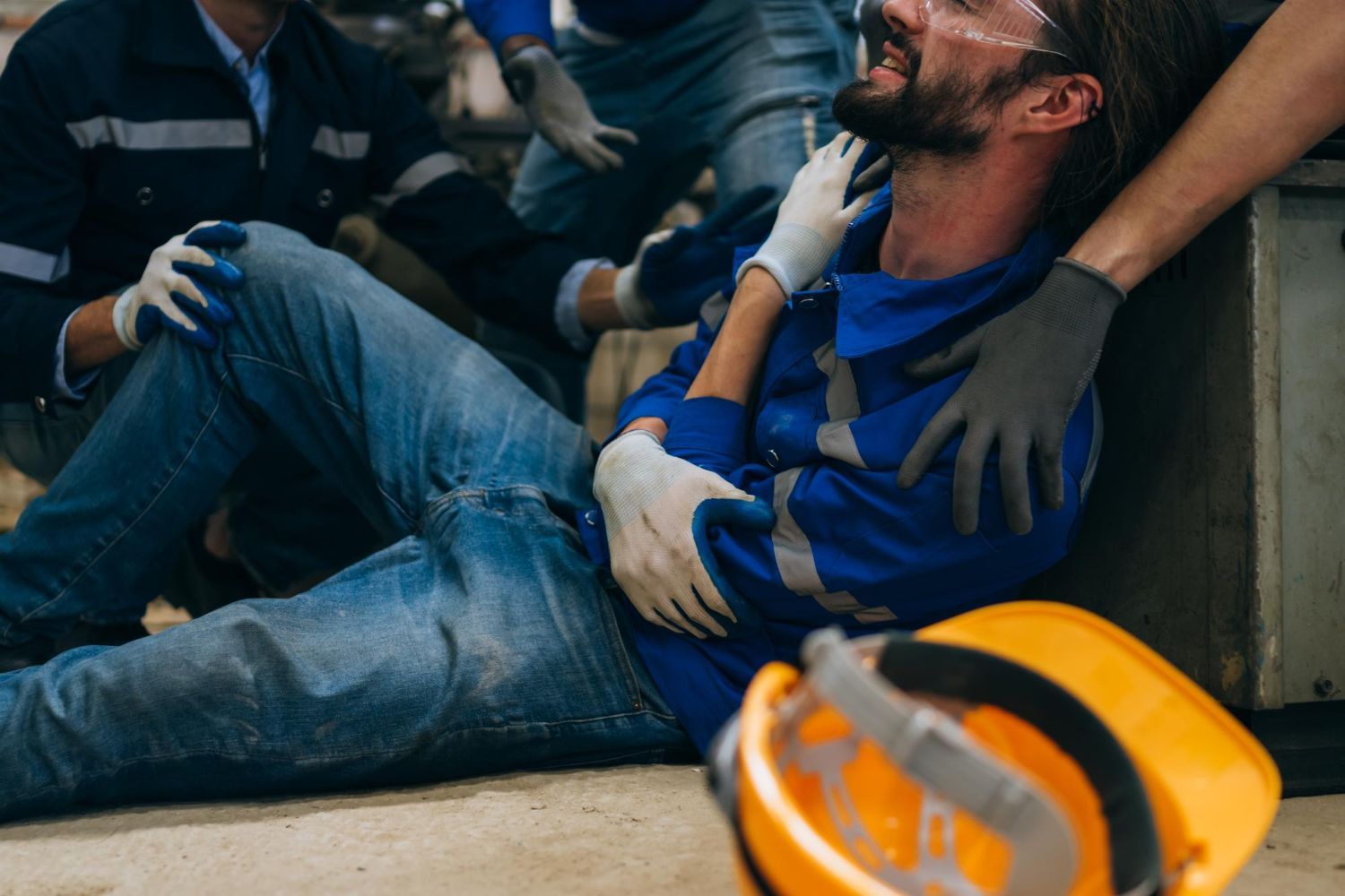 Man in blue work suit injured on floor, being helped by coworkers. Industrial setting.