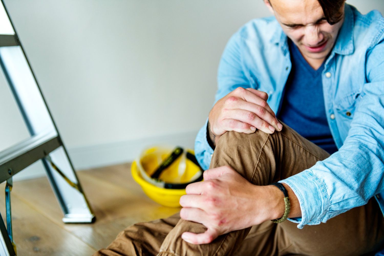 Man clutching his knee in pain after a fall, next to a ladder and a yellow hard hat.