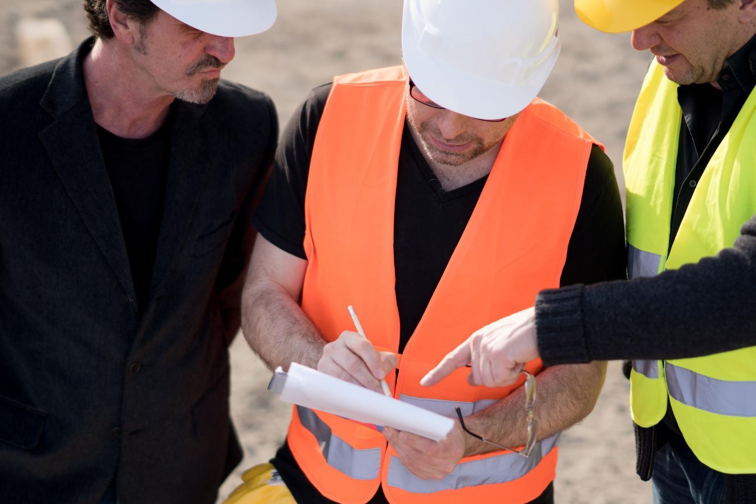 Three people, construction workers, reviewing blueprints on a construction site, one signing with a pen, all wearing hard hats and safety vests.