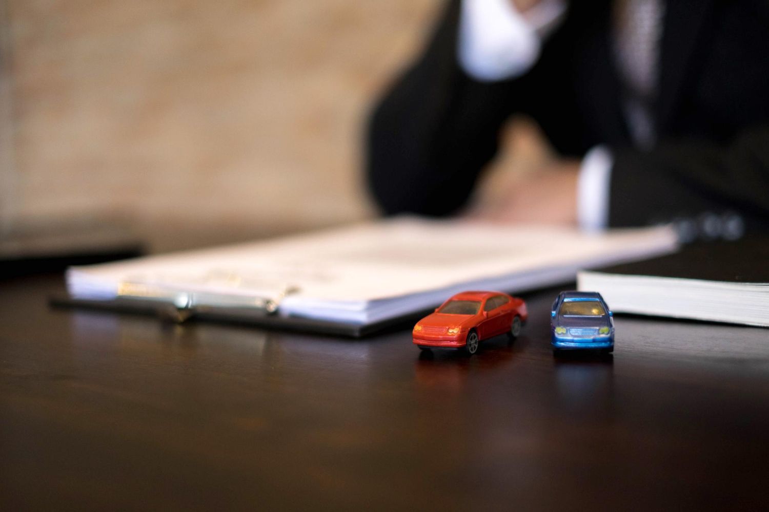 Miniature orange and blue cars on a desk with documents and a person in a suit, possibly insurance related.