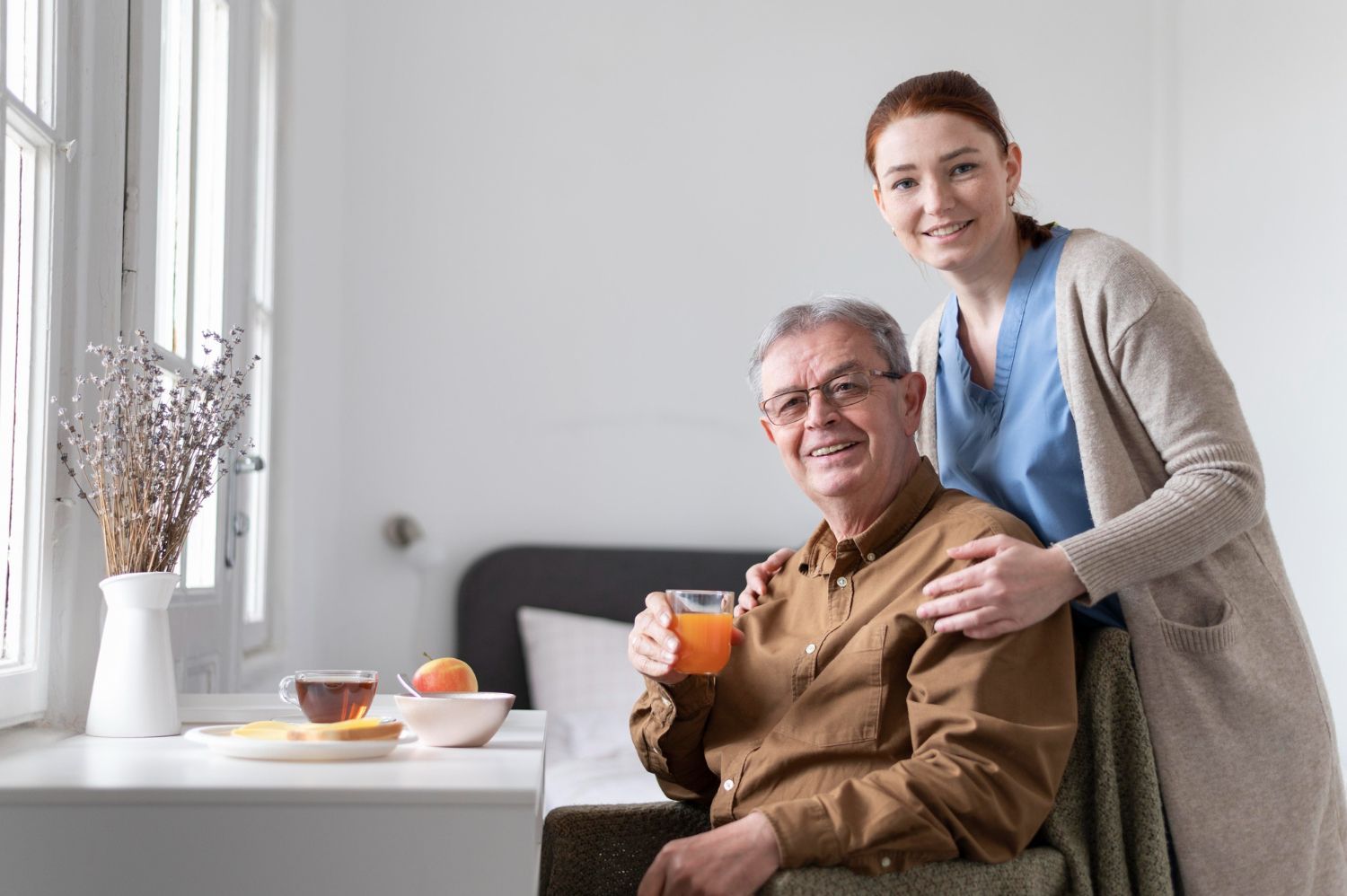 Senior man and caregiver smiling; he holds juice, breakfast on the table.