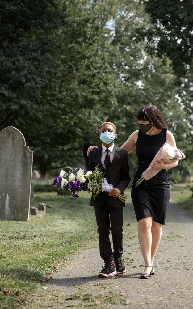 Woman in black dress comforts boy in suit, both wearing masks, walking in a cemetery, holding flowers.