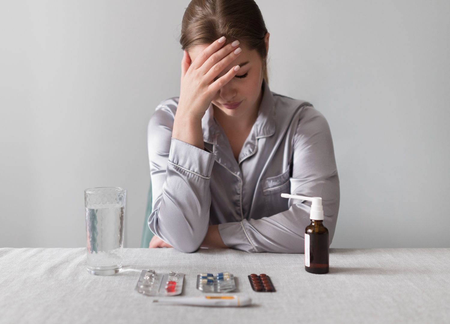 Woman with a headache sits at a table with medicine and a glass of water, looking unwell.