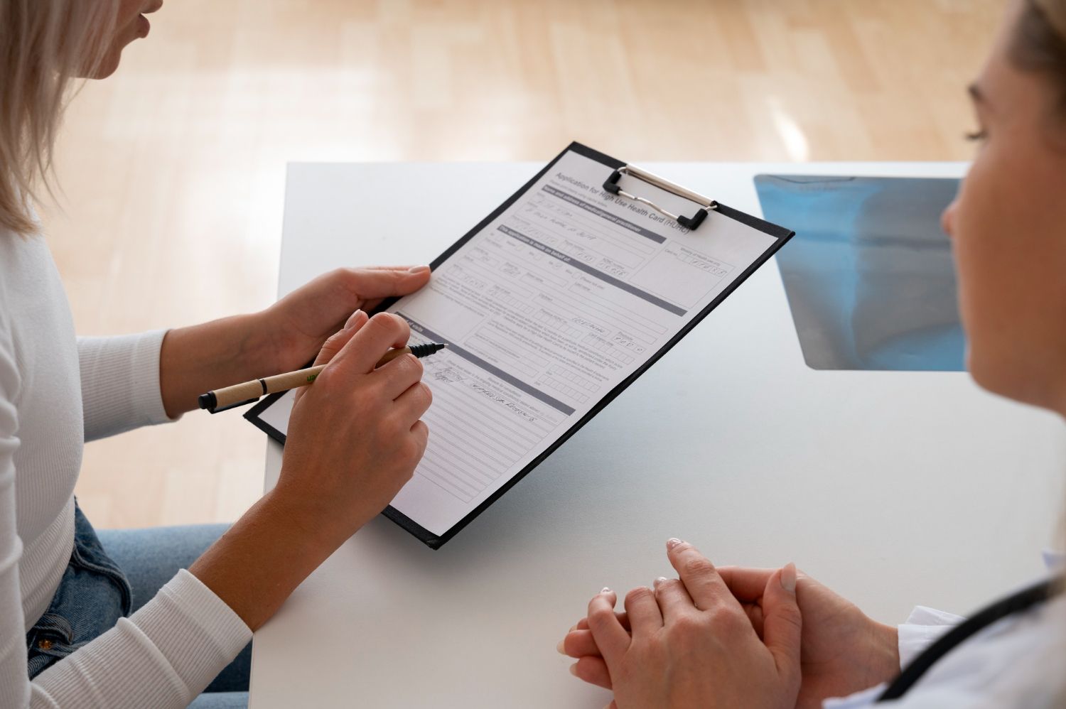Woman filling out paperwork, seated across from another woman, in a medical setting.