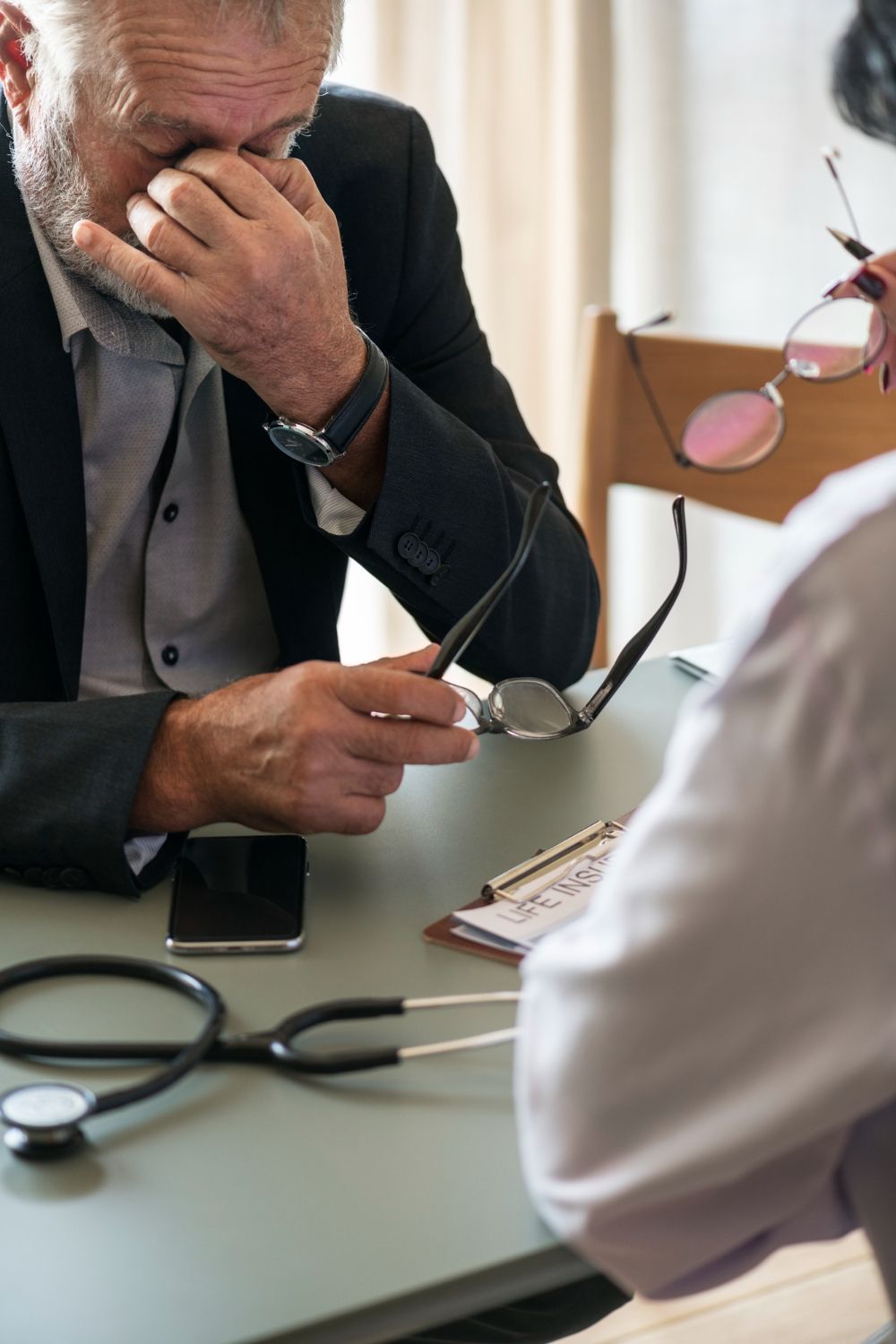 A man, distressed, covers his eyes in a doctor's office; stethoscope and notes on the table.