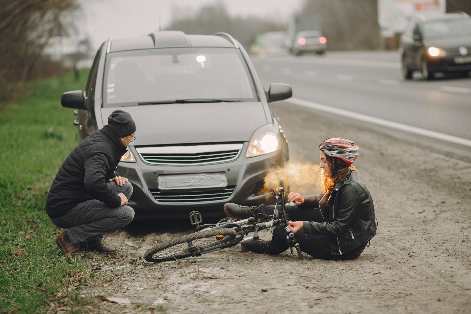 A cyclist sits injured on the ground next to a car; a man kneels beside them on a roadside.