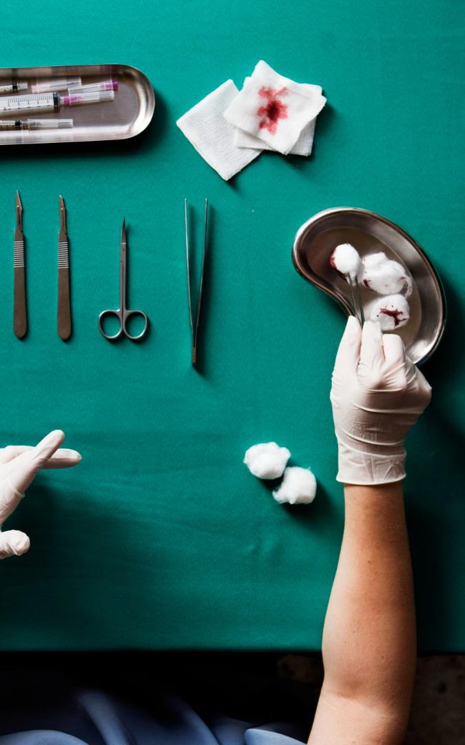 Surgical instruments and gloved hands on a green operating table. A tray holds bloody gauze, cotton balls, and a tray with tools.