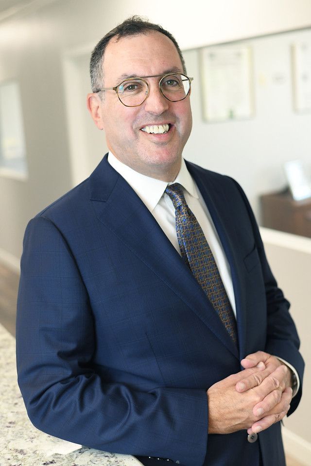 Man in a blue suit and glasses smiling, standing indoors.