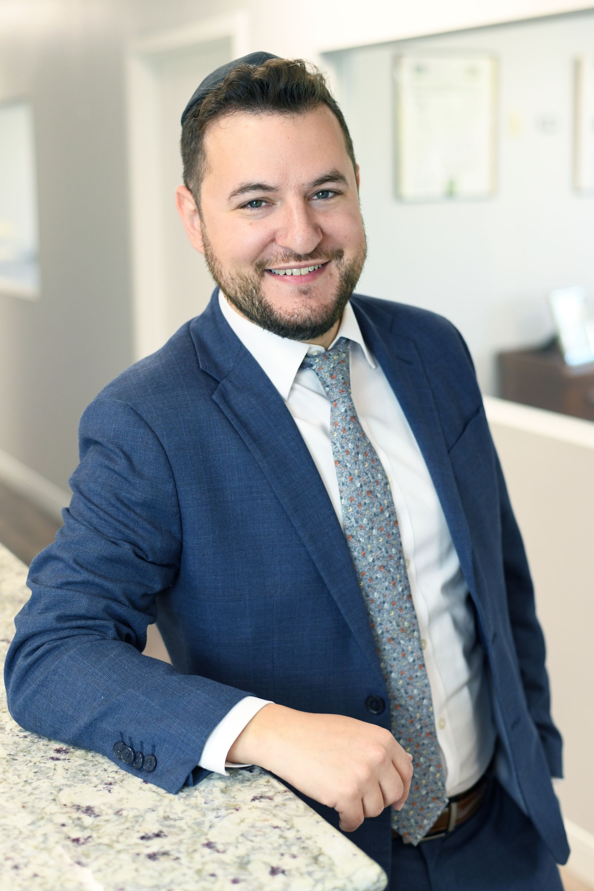 Man in a suit smiles, leaning on a counter. He has gray hair and a green tie.