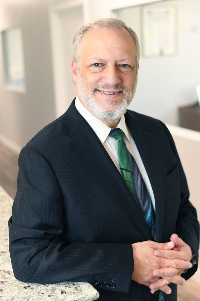 Man in a suit smiles, leaning on a counter. He has gray hair and a green tie.