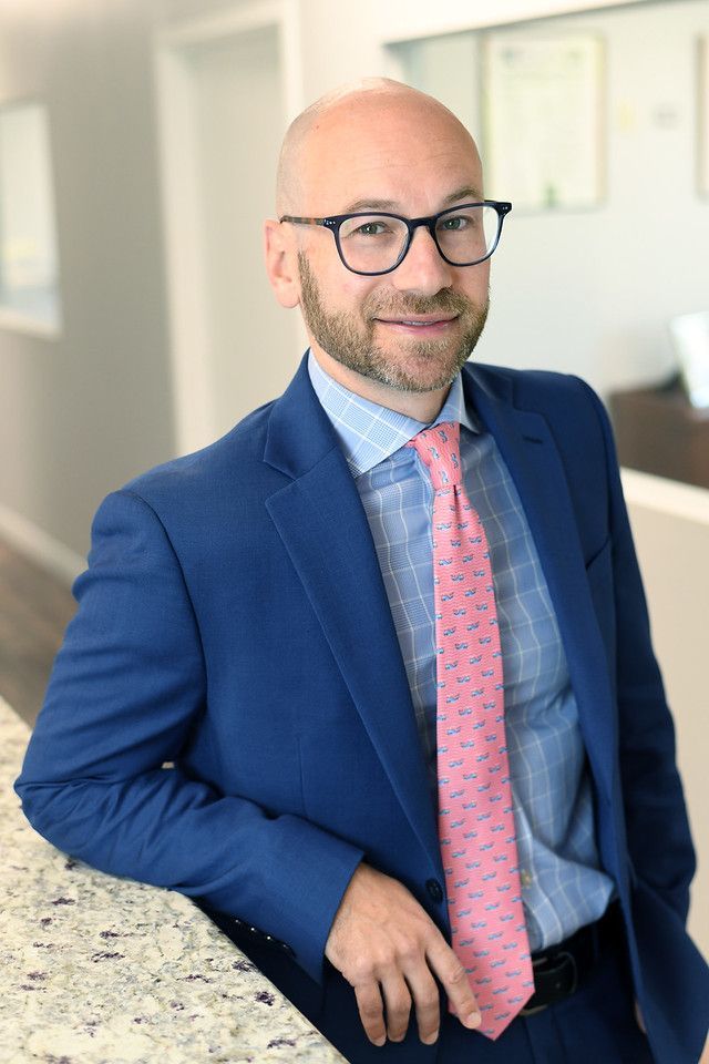 Man in blue suit and glasses, smiling, leaning on a counter. Pink tie, light-colored shirt. Indoor, professional setting.