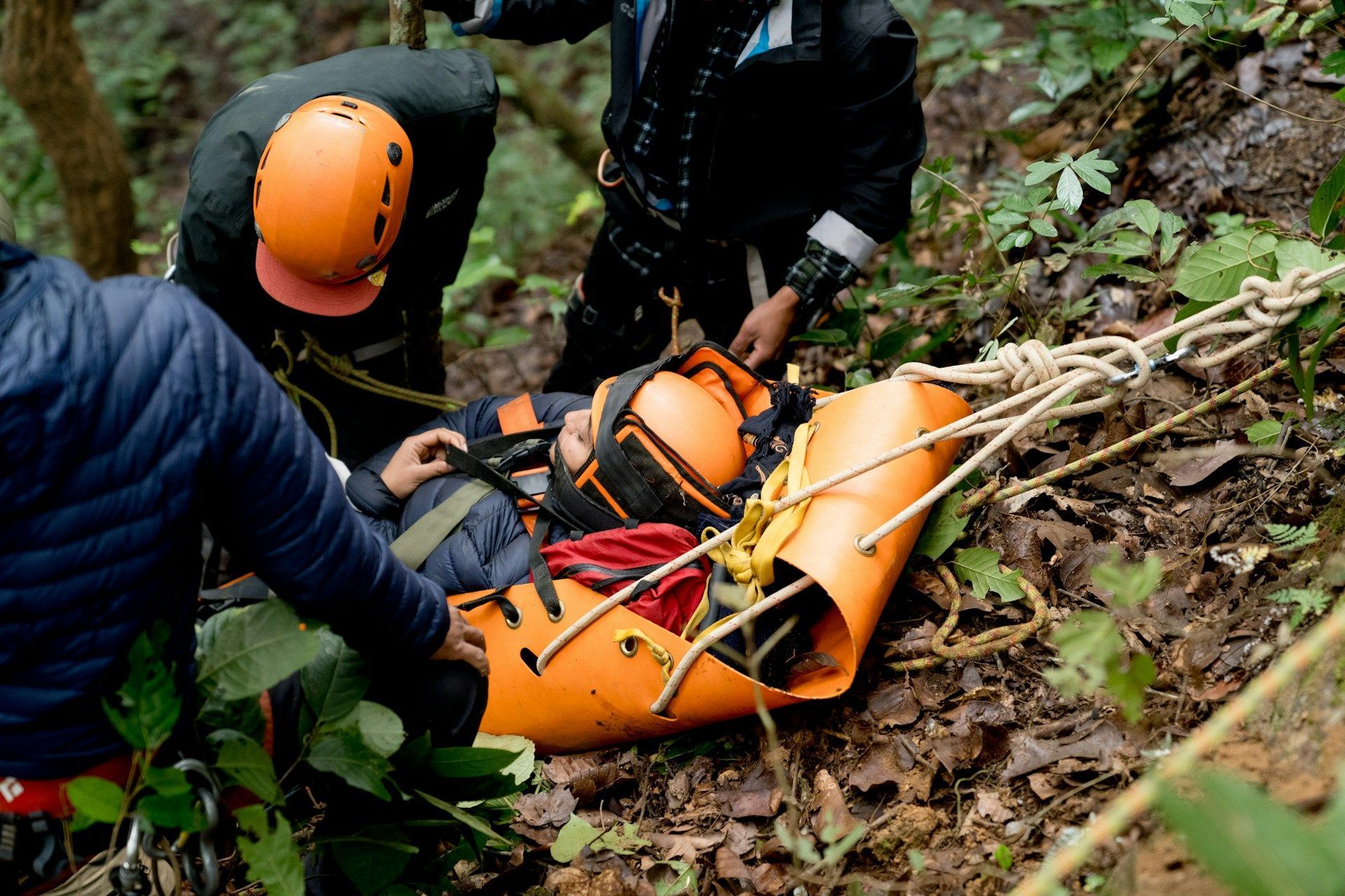 Rescue workers securing an injured person in an orange stretcher on a wooded hillside.