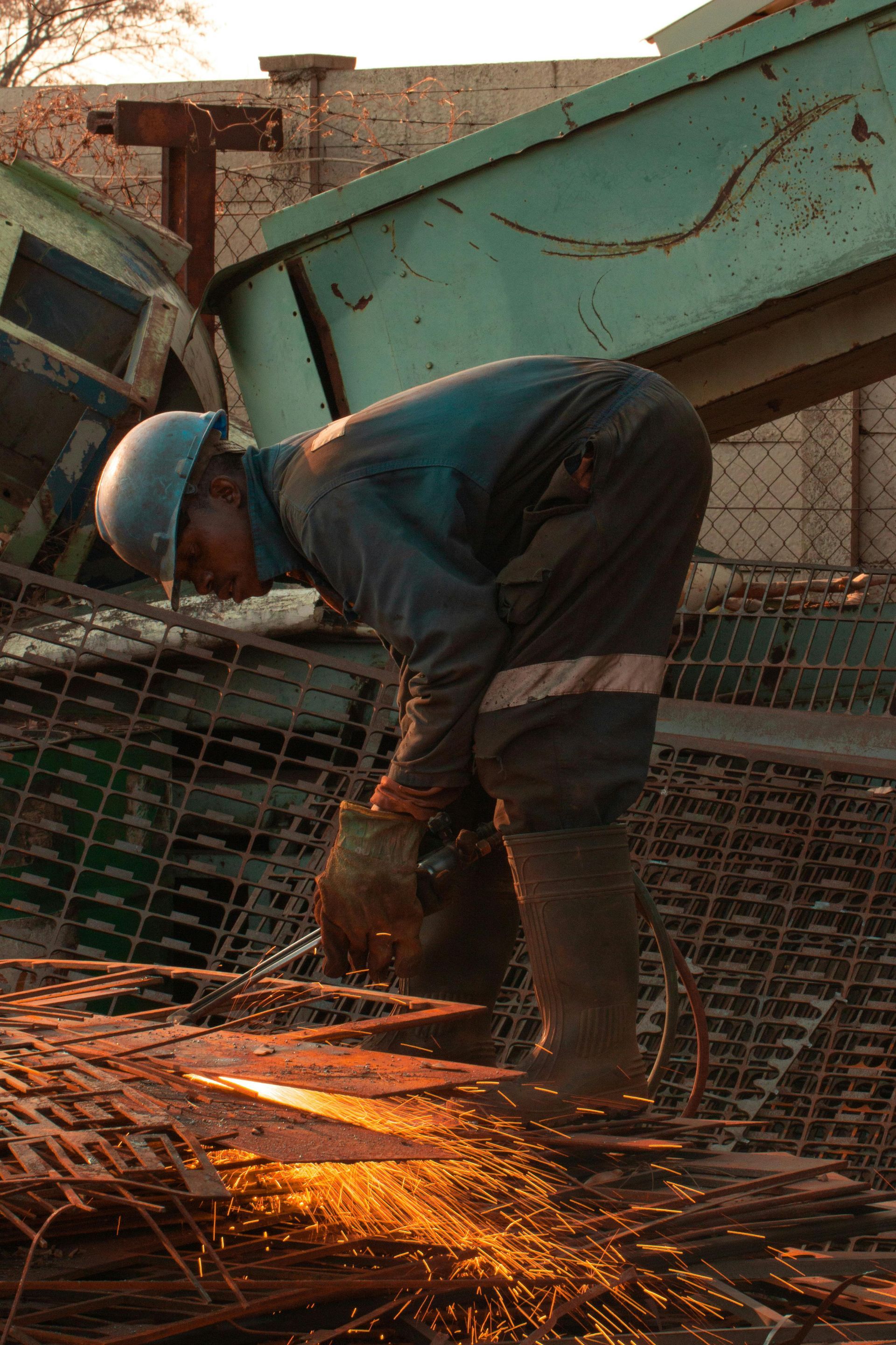 Man in work clothes using a grinder, sparks flying; rusty metal and debris in a sunlit area.
