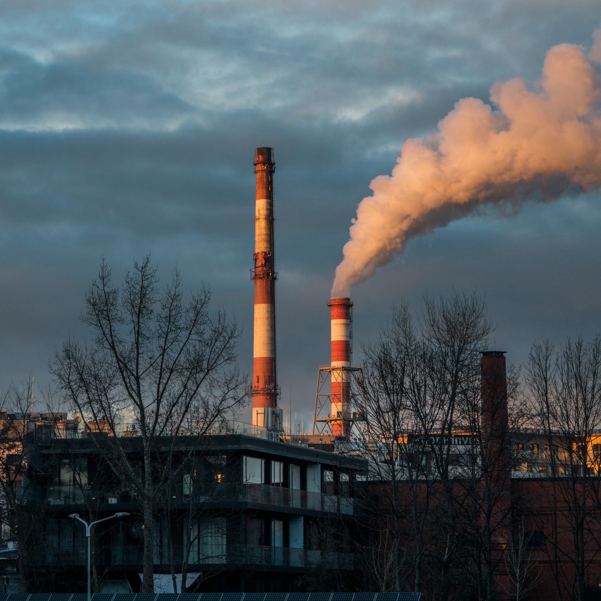 Factory chimneys spew white smoke into a cloudy sky. Buildings and bare trees in foreground.