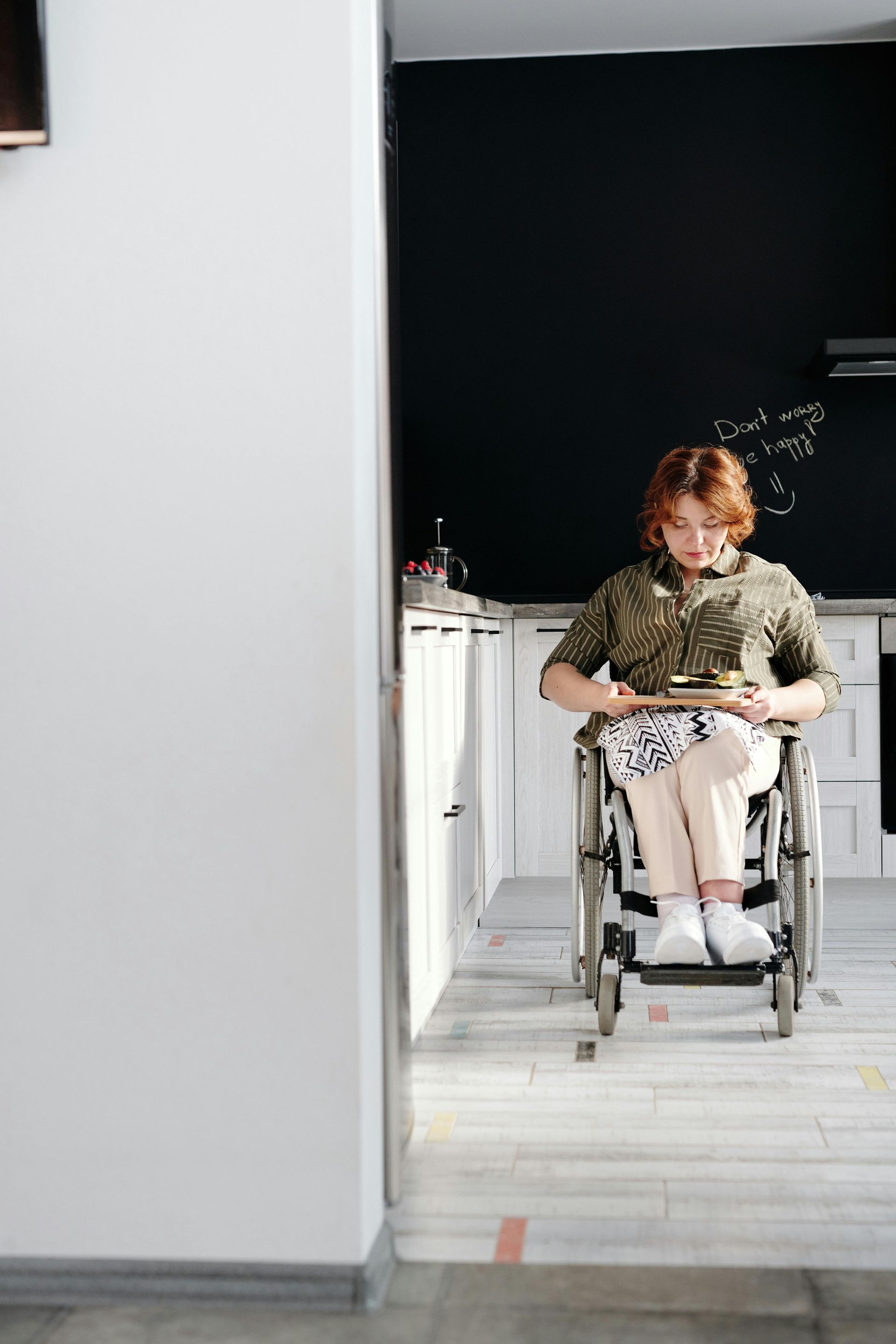 Person in wheelchair holding a small plate in a kitchen. They have red hair, are wearing patterned clothing, and are facing forward.