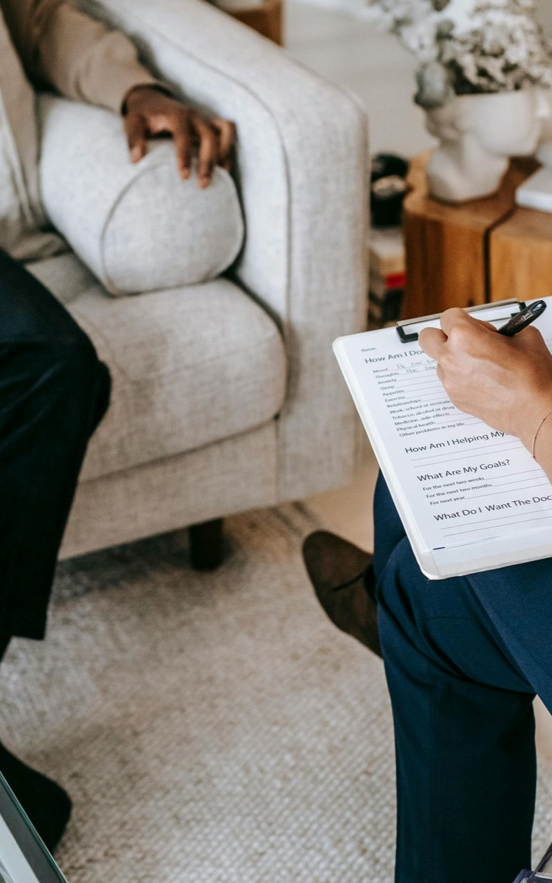 Therapist taking notes on a clipboard while the client sits in an armchair, indoor setting.