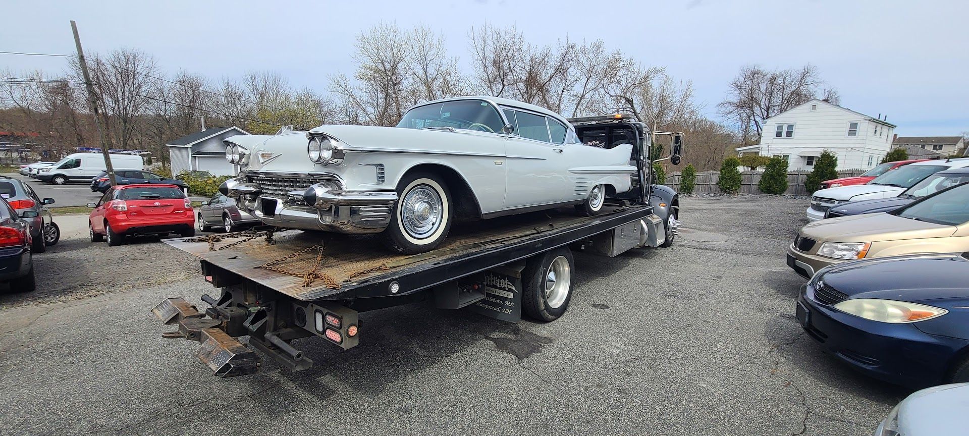 A classic white car on a tow truck in a gravel lot with other cars.