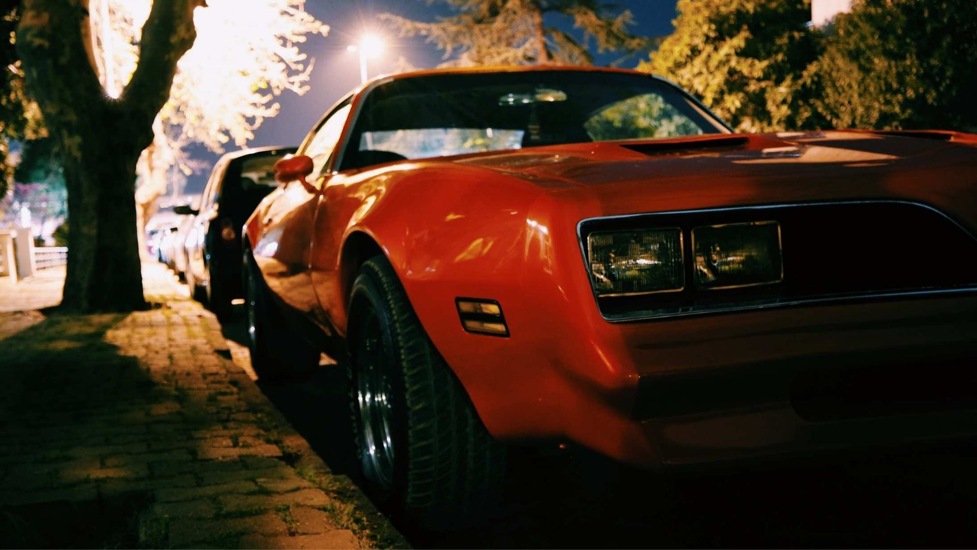 Red classic car parked on a tree-lined street at night, illuminated by streetlights.