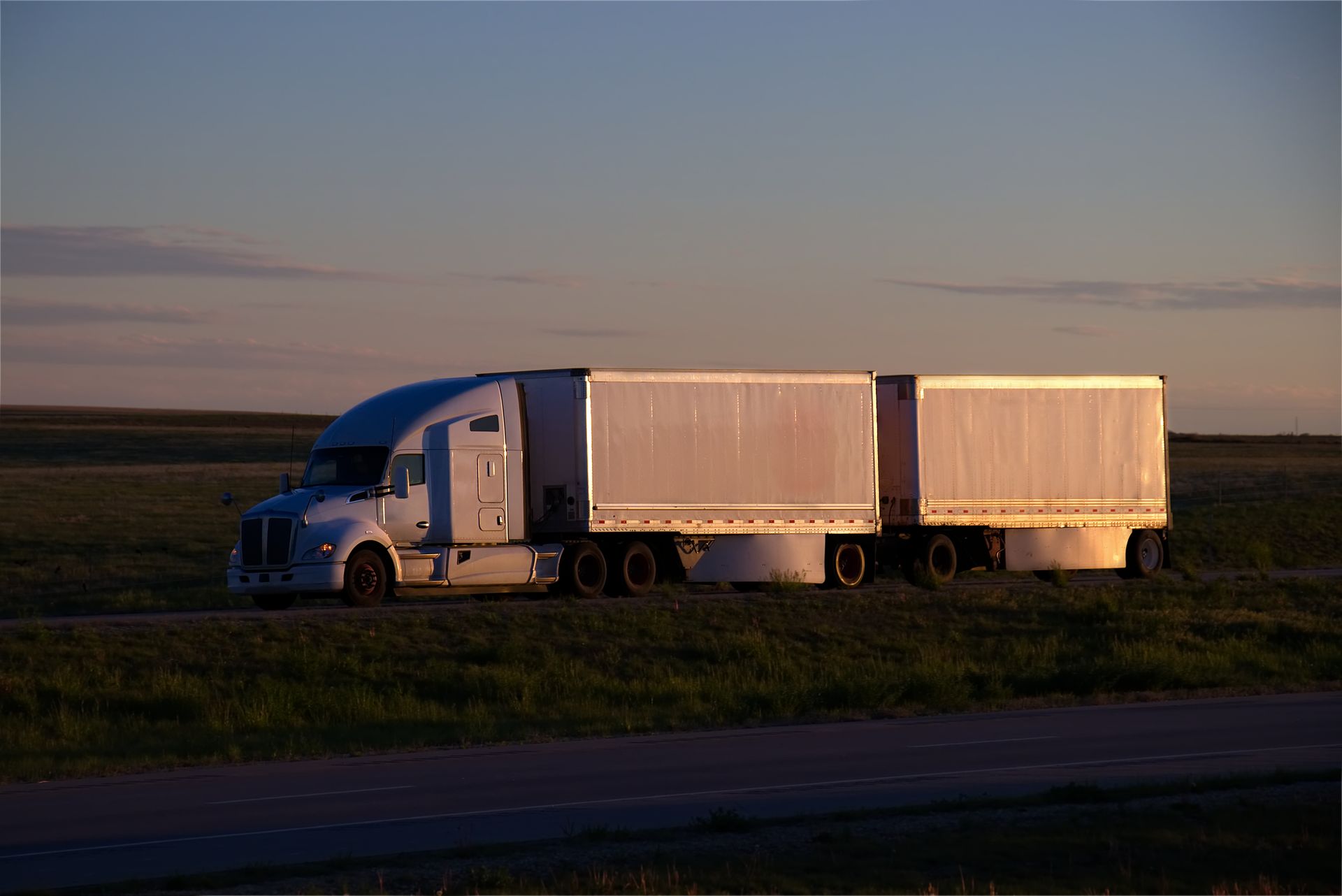 White semi-truck with two trailers on a road in a grassy field at sunset.