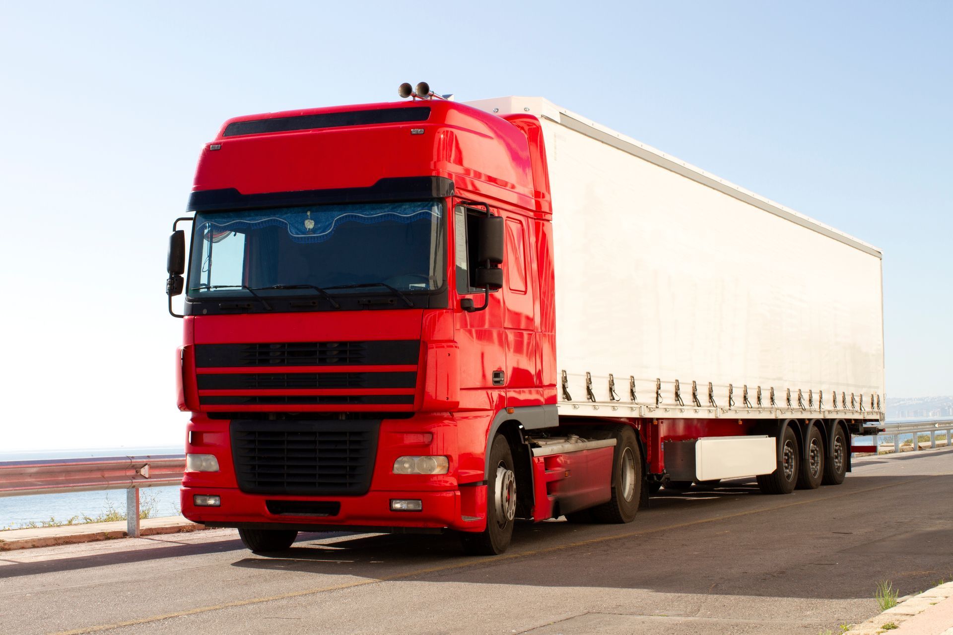 Red semi-truck with a white trailer driving on a road under a blue sky.