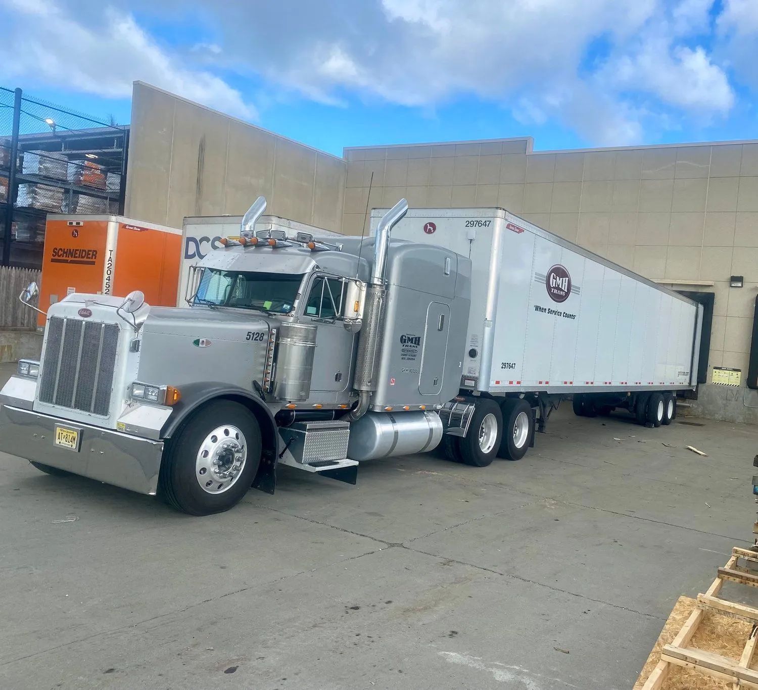 Silver semi-truck with trailer parked at loading dock. Sky is blue with clouds.