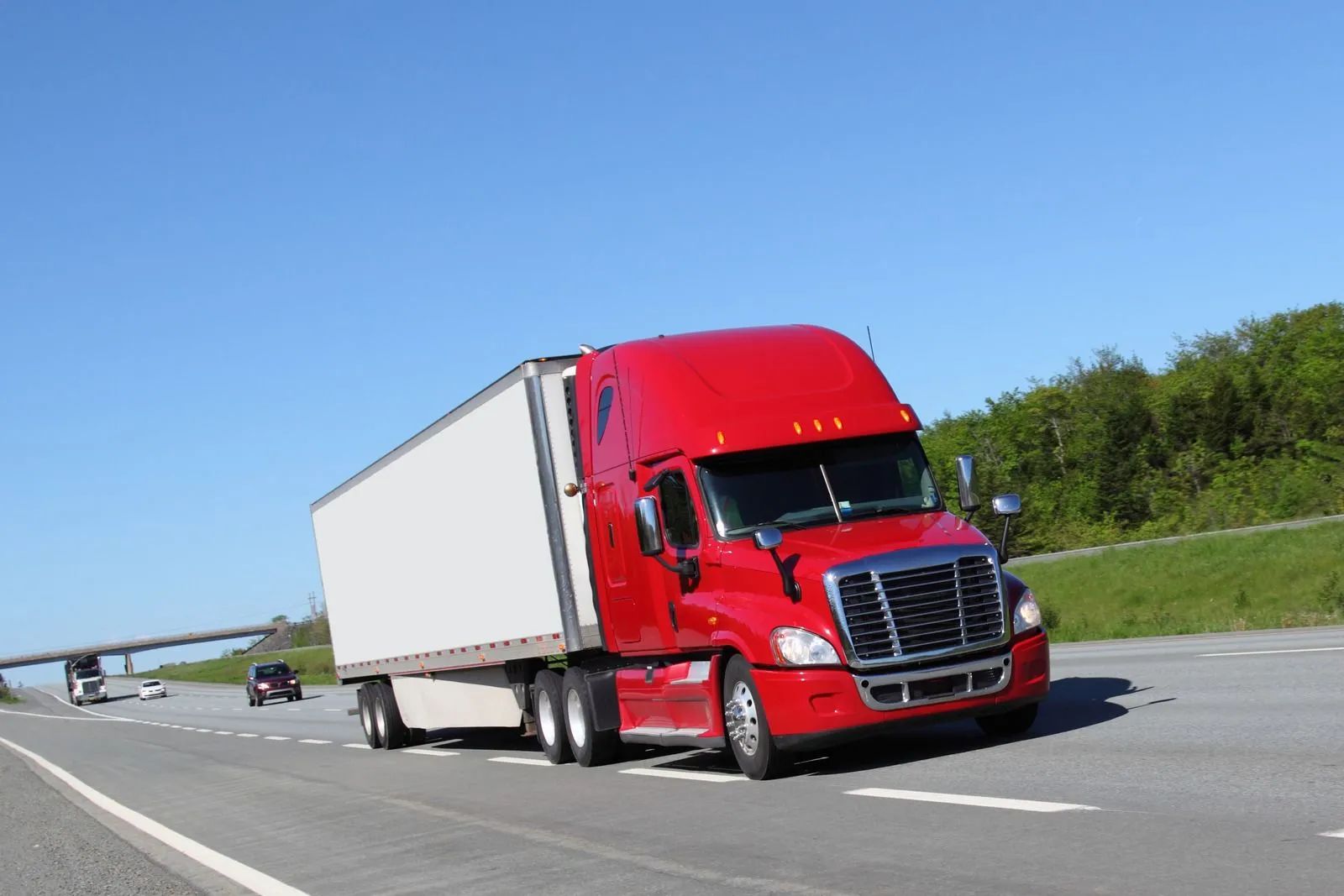 A red semi-truck with a white trailer drives down a multi-lane highway on a clear, sunny day.
