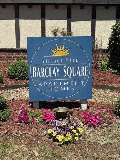 Sign for Barclay Square Apartment Homes, blue with gold sun and text, surrounded by flowers.