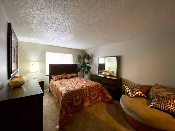 Bedroom with a bed, dresser, mirror, and curved sofa. Warm color tones dominate.