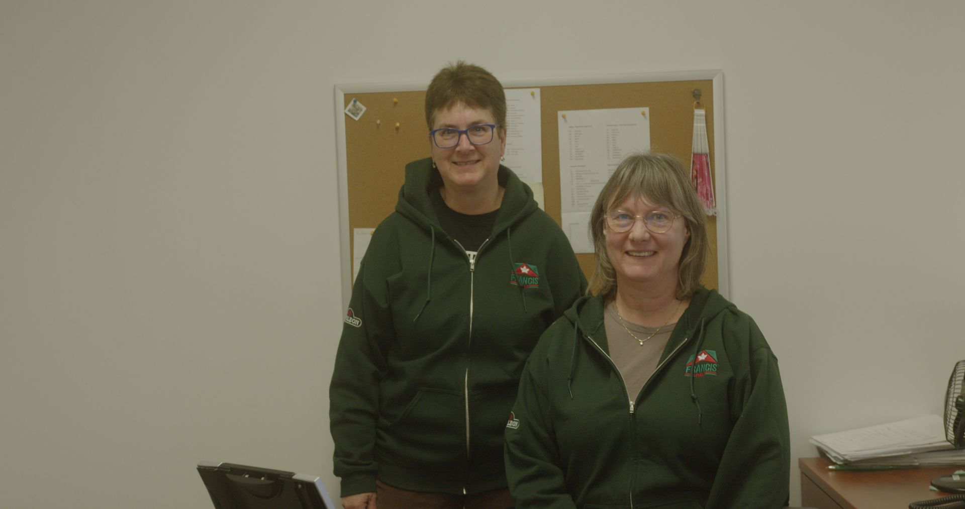 Two women are standing next to each other in front of a bulletin board.