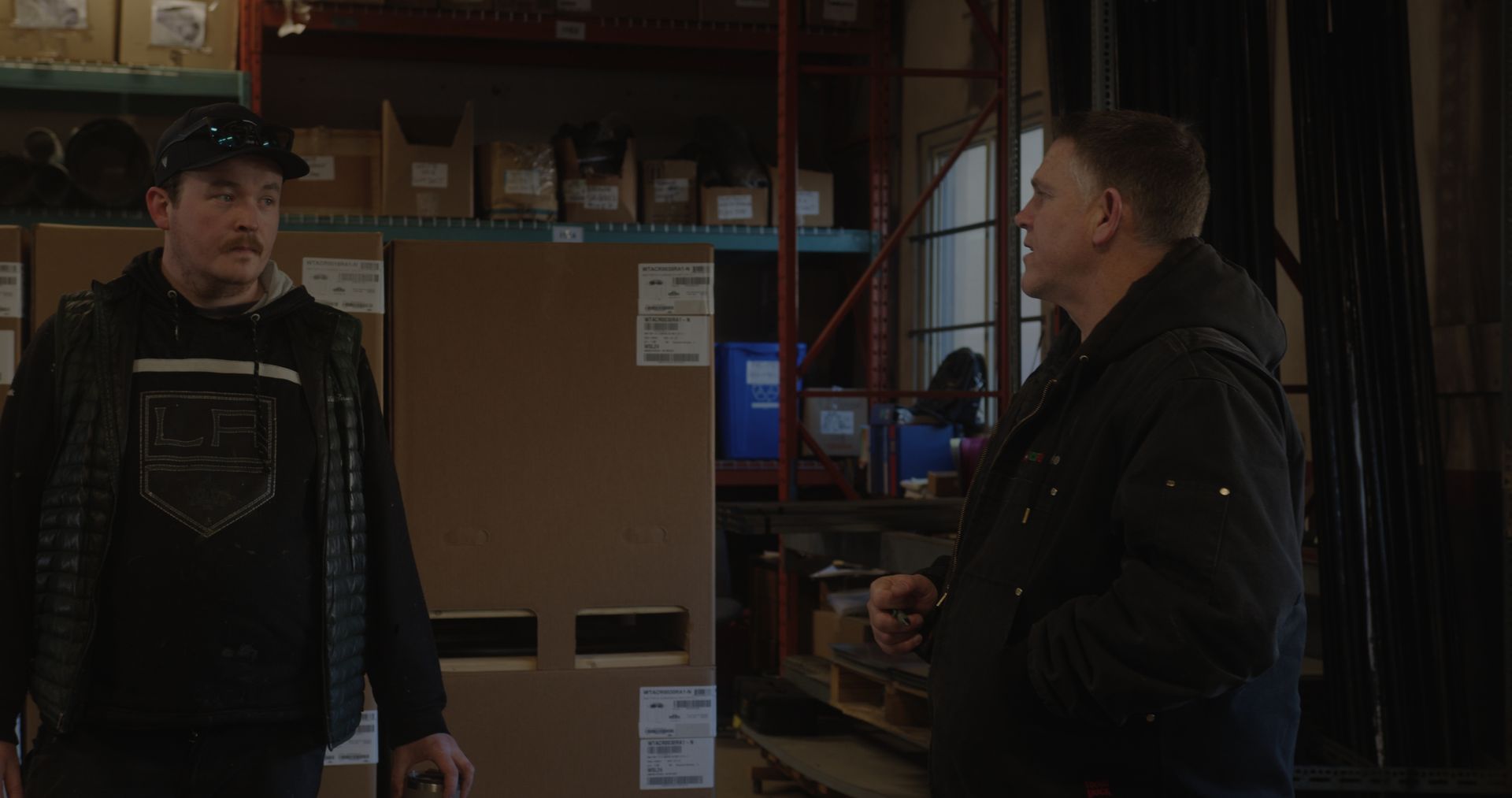 Two men are standing in a warehouse talking to each other.