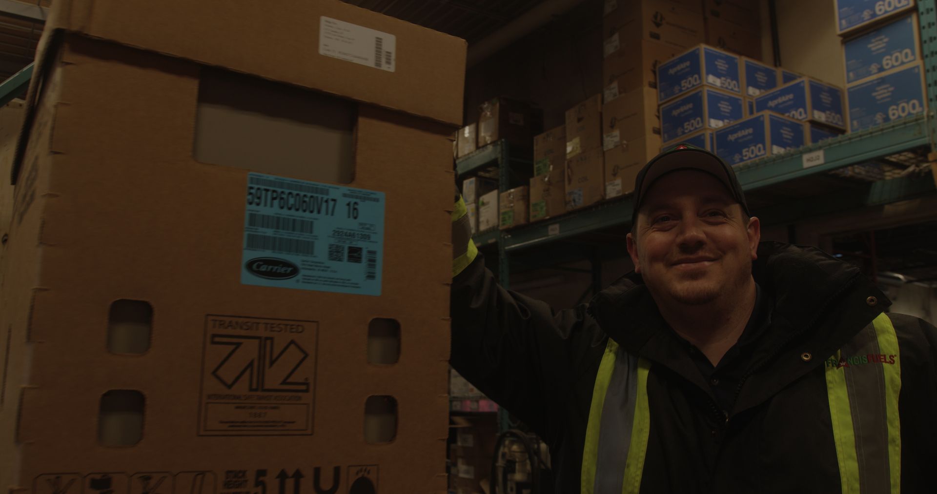 A man is standing in front of a large cardboard box in a warehouse.